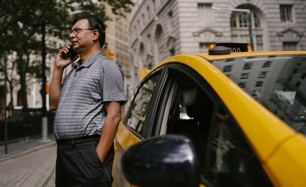 Serious ethnic male taxi driver with hand in pocket having conversation on cellphone while standing near car